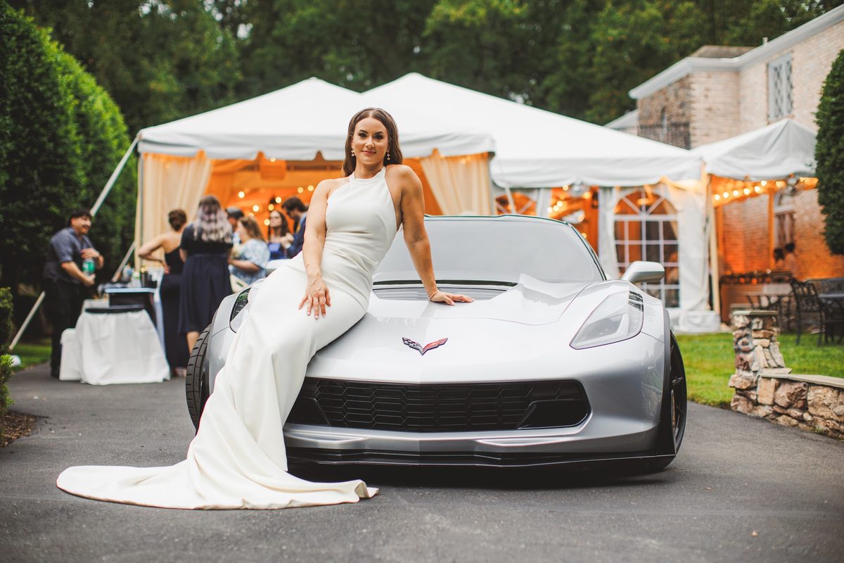Bailey seated on the hood of a silver Corvette in the driveway, her long train pooling to the asphalt, the reception tent glowing amber behind her