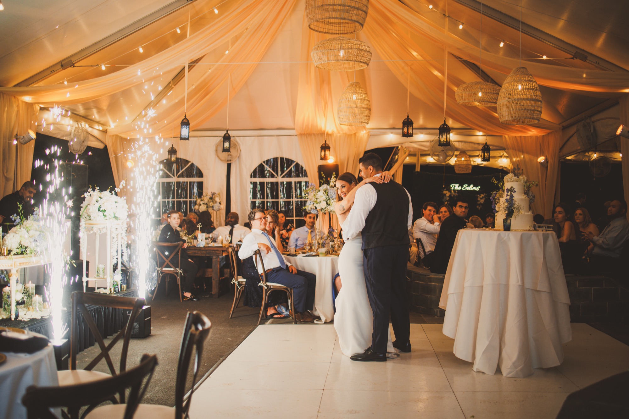 Bailey and Youssef dancing their first dance under a warmly lit reception tent as spark fountains fire beside them, guests watching from tables and a tiered cake at the edge of the floor