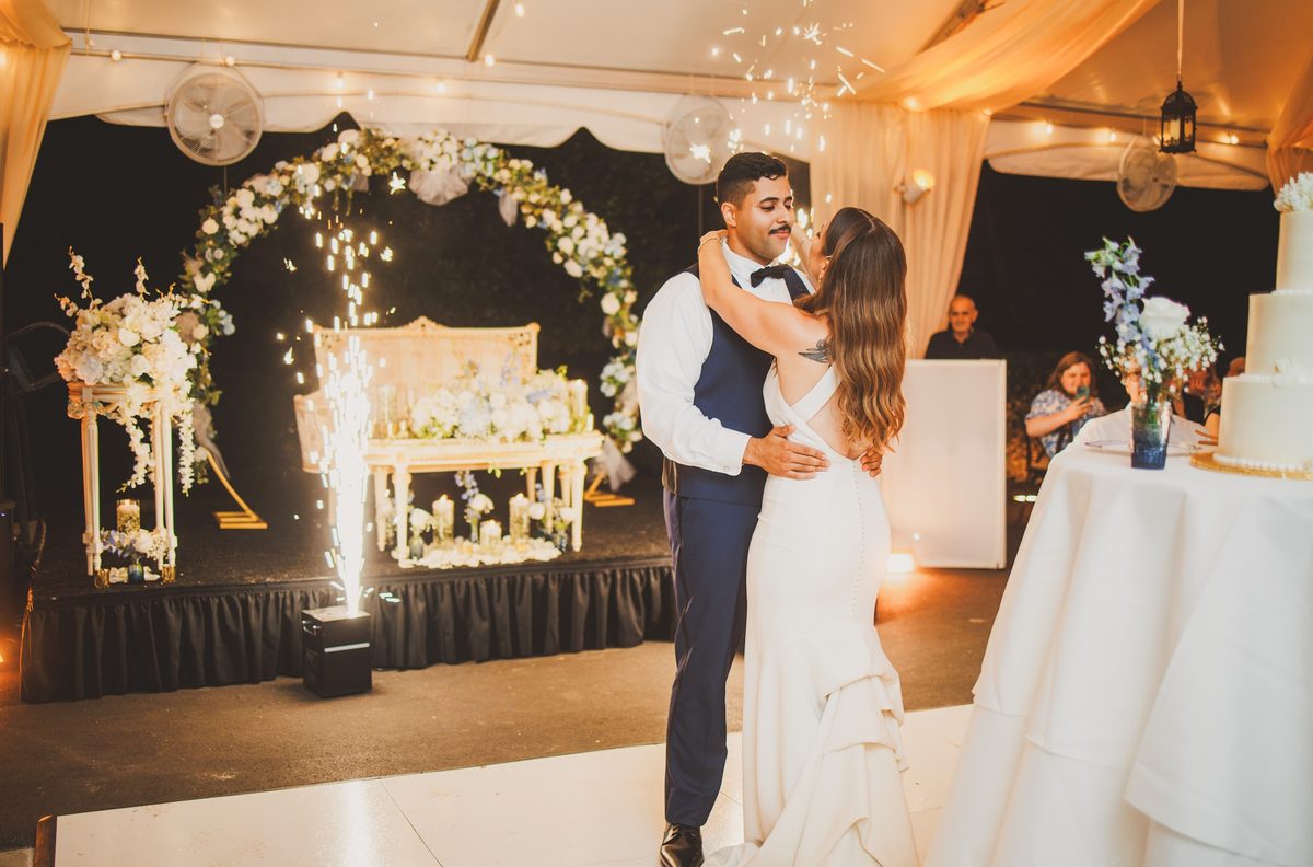 Bailey and Youssef slow dancing on the white dance floor as cold-spark fountains rise beside the flower-arched sweetheart stage