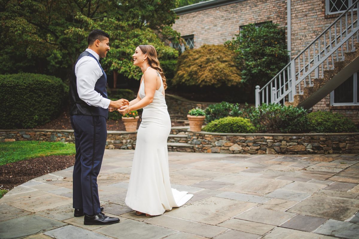 Youssef holding both of Bailey's hands on the flagstone patio during the first look, both laughing, stone walls and greenery behind