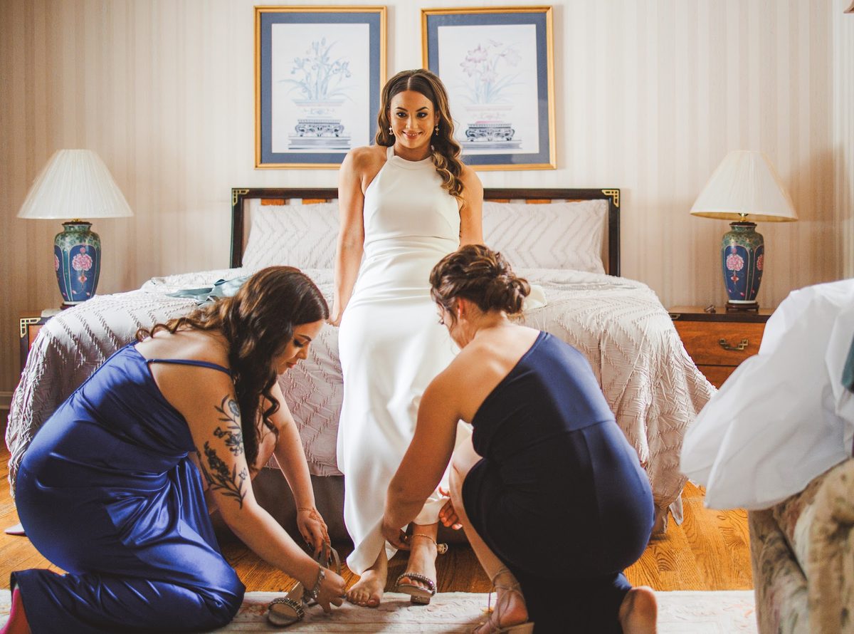 Bailey in her white halter crepe gown perched on the edge of the bed while two bridesmaids in navy dresses fasten her sparkling heels