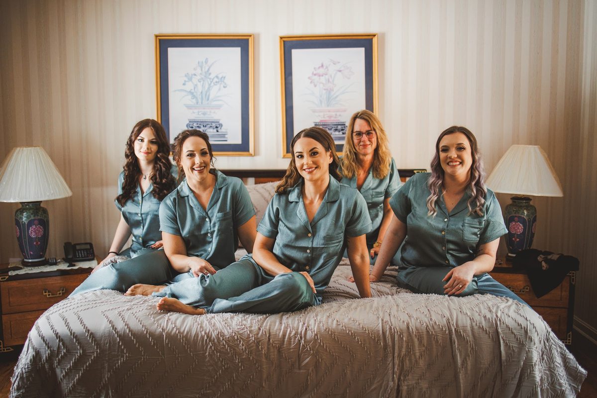 Bailey and four of her bridal party in matching sage green silk pajamas sitting together on a bed between two lamps