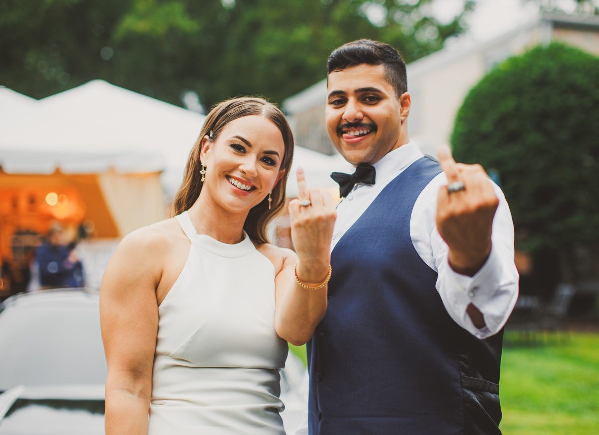 Bailey and Youssef grinning at the camera and holding up their ring fingers, new bands on display, the glowing tent behind them