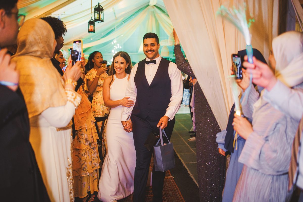 Bailey and Youssef walking out through guests waving glowing light wands and raising phones, both beaming