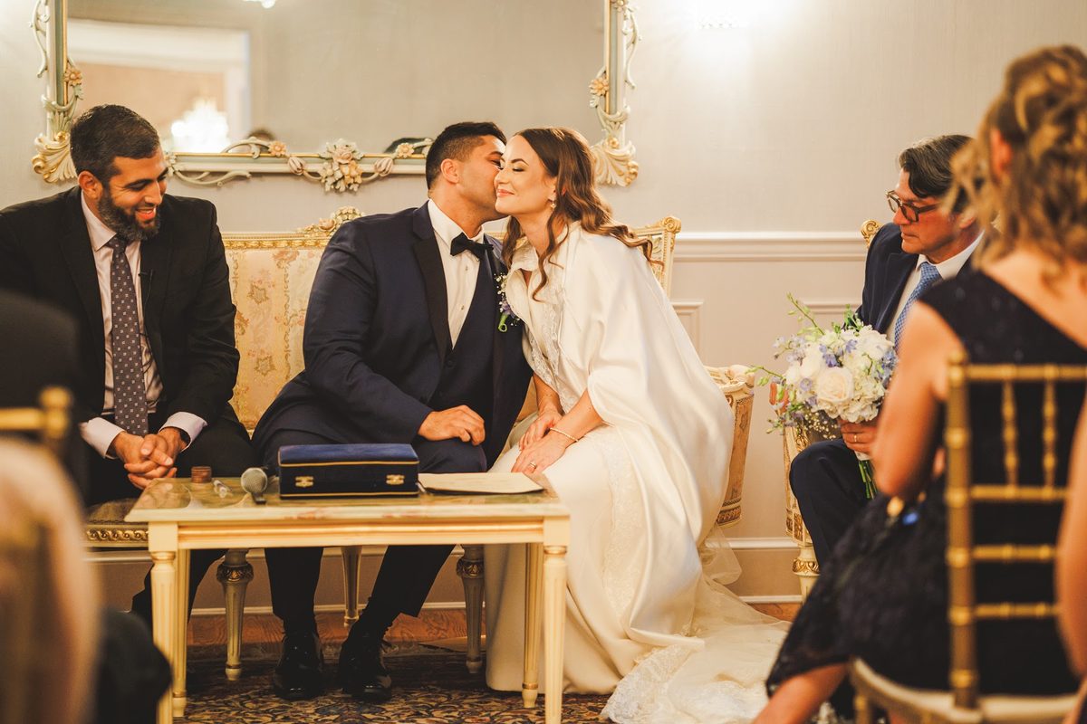 Youssef kissing Bailey's cheek at the signing table as she closes her eyes and smiles, the officiant and family smiling on either side