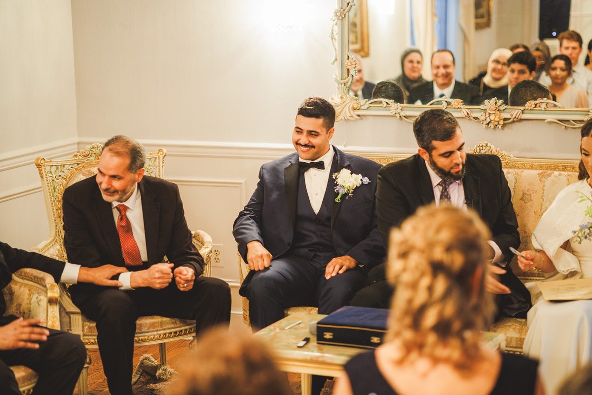 Youssef grinning in his navy tuxedo beside the officiant during the ceremony while Bailey signs the marriage document at the edge of the frame, family reflected in the mirror above