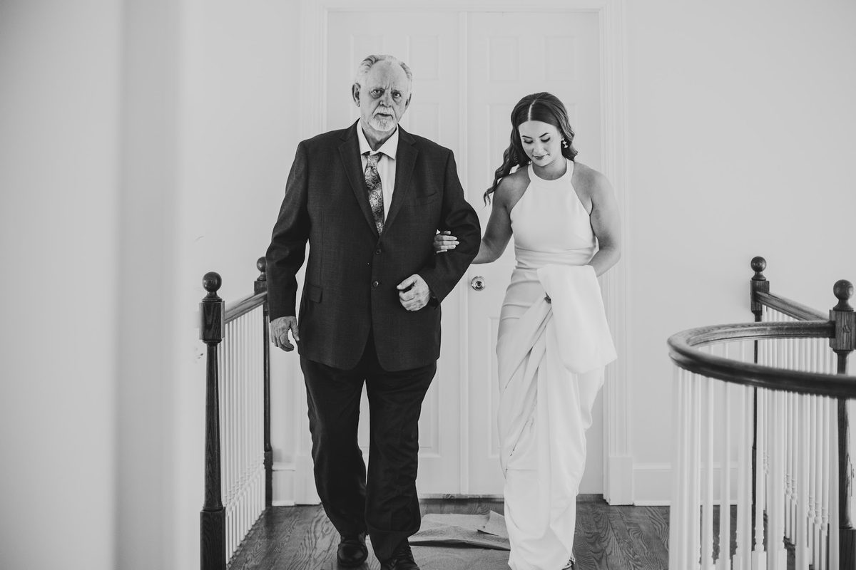 Black and white frame of Bailey on her grandfather's arm at the top of the staircase, holding her gathered train, about to walk down
