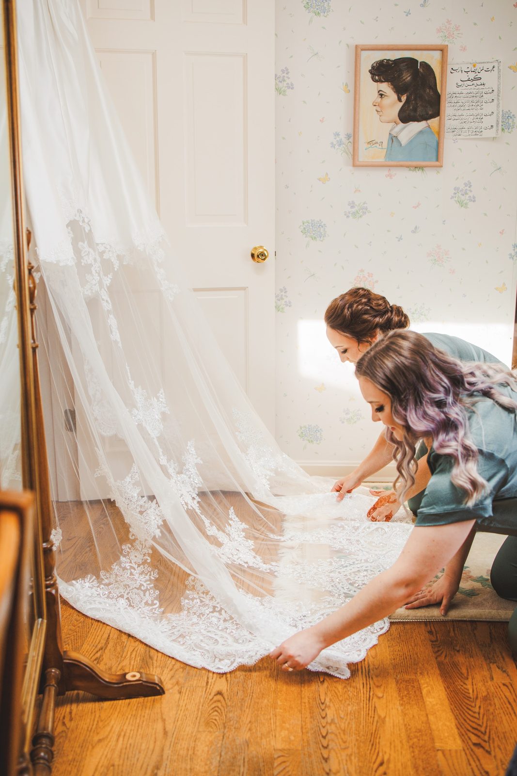 Two bridesmaids in sage green pajama sets smoothing Bailey's long lace-edged veil across the wood floor by a bright window