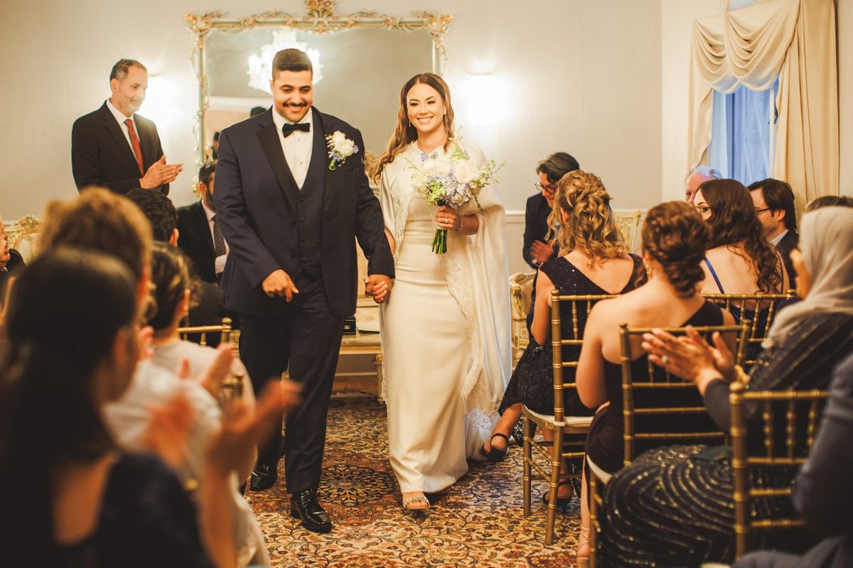 Bailey and Youssef walking hand in hand up the rug-lined aisle between applauding guests after the ceremony, her bouquet in hand