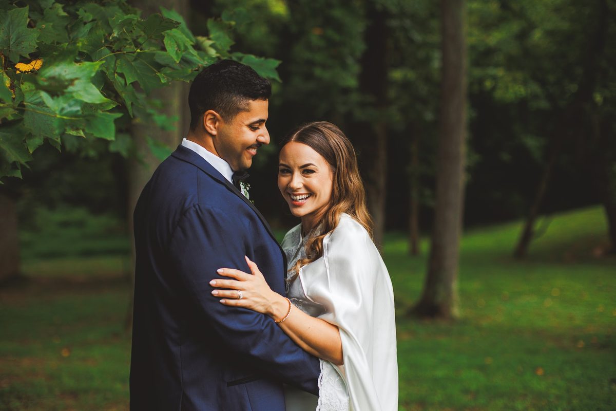 Bailey and Youssef nose to nose beneath towering trees at a Brinklow, Maryland estate, her white satin cape and lace-edged train spread across the grass