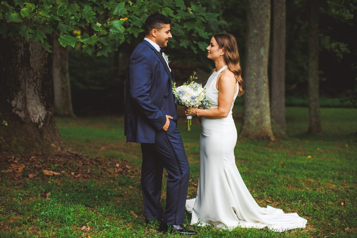 Bailey and Youssef facing each other among the tall tree trunks, her bouquet of white and pale blue held between them