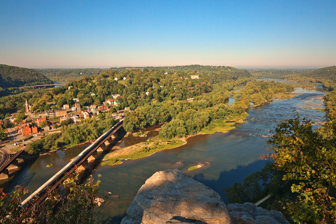 Harpers Ferry seen from the heights above, the town stacked on the point where the Shenandoah meets the Potomac, the railroad bridge crossing below and wooded ridges running off into the distance