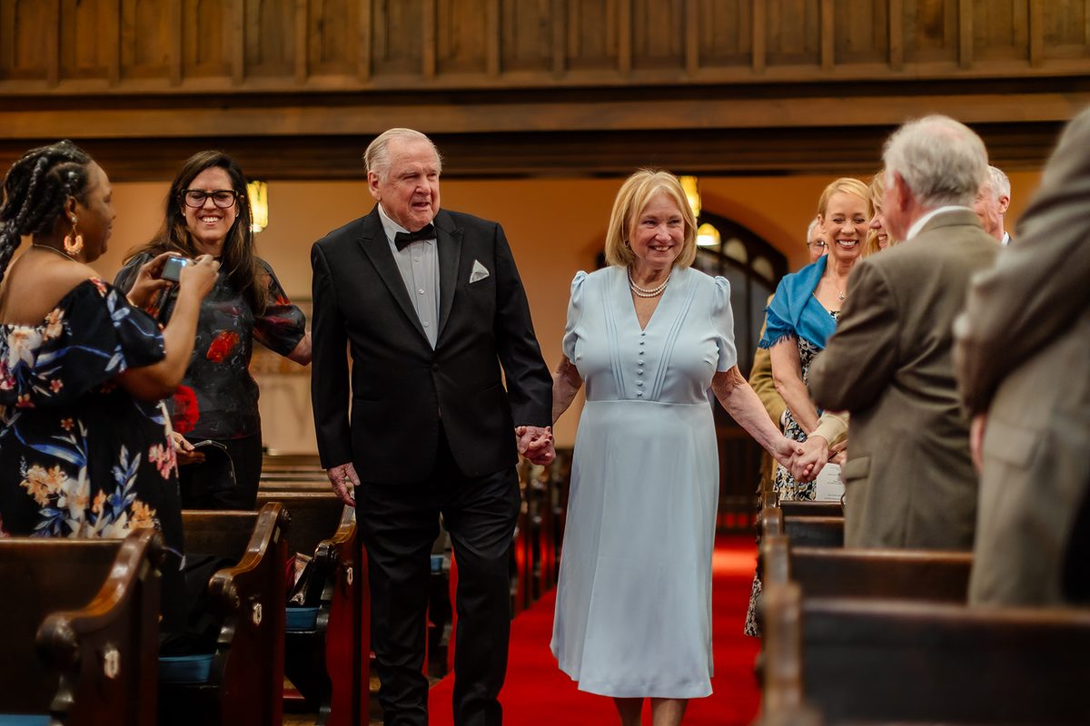 Mary in her pale blue dress and Mark in his tuxedo walking up the red-carpeted aisle side by side, smiling at their guests