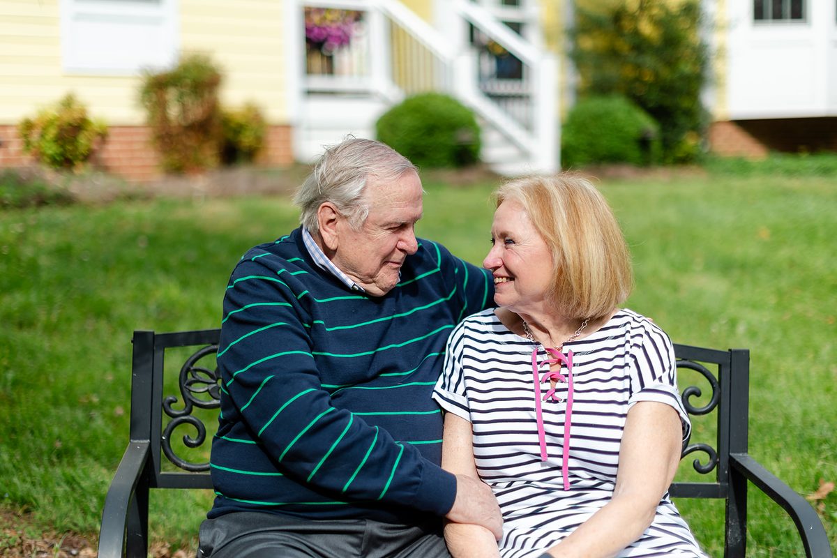 Mary and Mark sitting close together on a garden bench in front of a yellow house, her hand resting on his chest as they look at each other