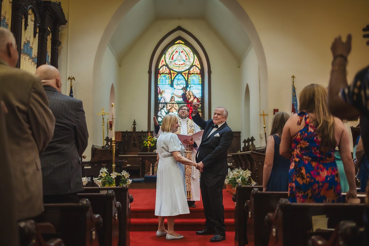 Mark raising a fist in celebration at the chancel as Mary laughs beside him and the priest smiles