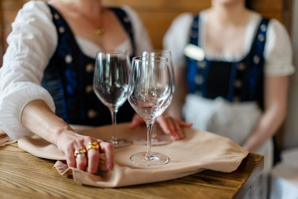 A Bavarian Inn server in a traditional dirndl setting rows of wine glasses in the bright reception room