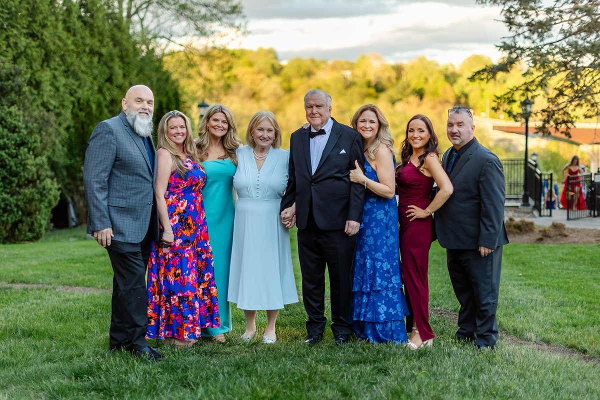 Mary and Mark surrounded by family in jewel-toned dresses and dark suits on the Bavarian Inn lawn, the treed valley glowing gold behind them