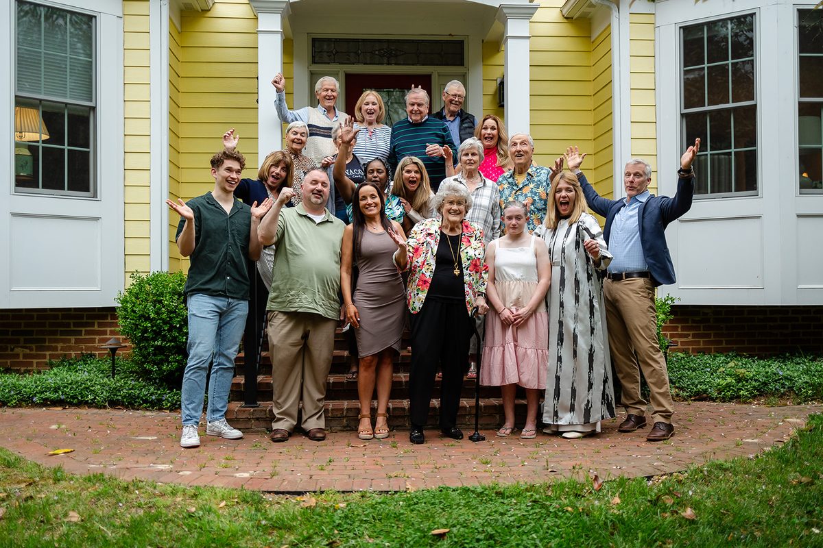 A large group of family and friends packed onto the front steps of the yellow house, arms in the air, everyone cheering at the camera
