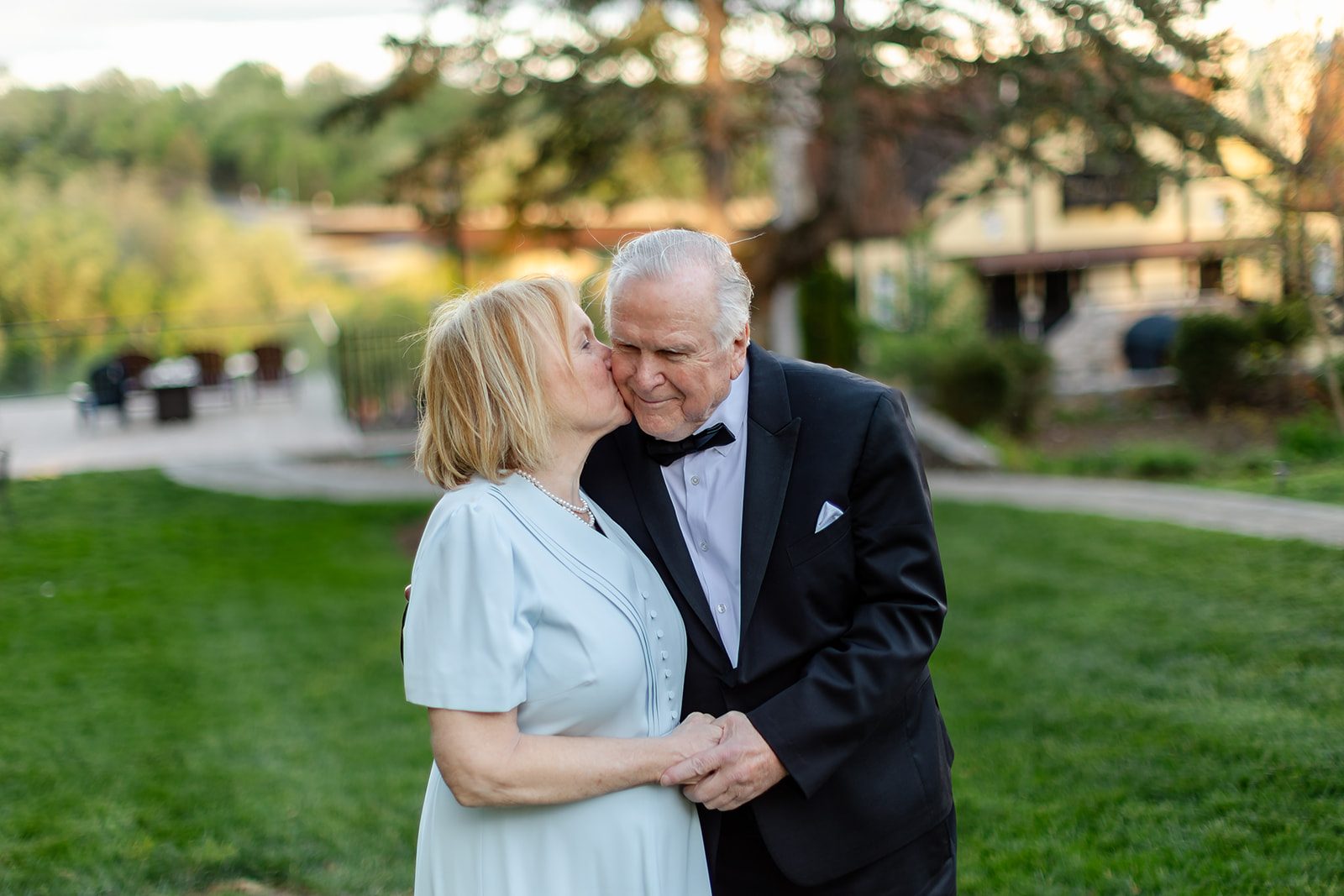 Mark kissing Mary on the cheek in golden evening light on the Bavarian Inn lawn, her flowers in hand and the treed river valley soft behind them