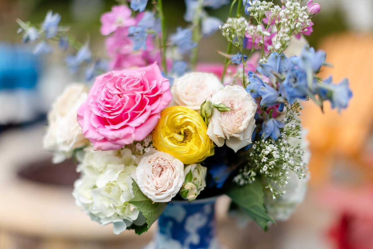 Close view of an arrangement of pink roses, a yellow ranunculus, blue delphinium, white hydrangea, and baby's breath in a blue and white vase