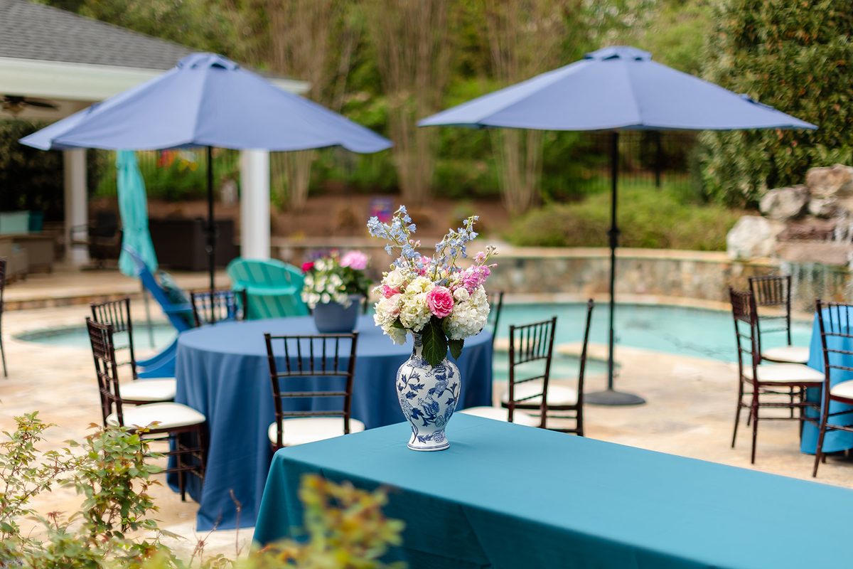 Round tables in blue and teal linens set around a stone pool with navy umbrellas, an arrangement of pink roses and blue delphinium in a blue and white vase in the foreground