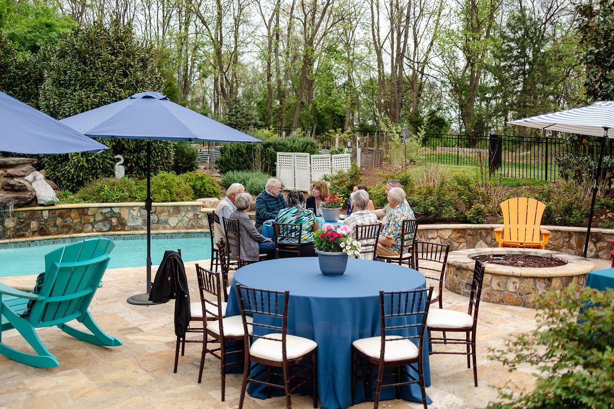 Guests gathered at a table by the pool under navy umbrellas, a fire pit and bright yellow and teal chairs around them, woods rising behind