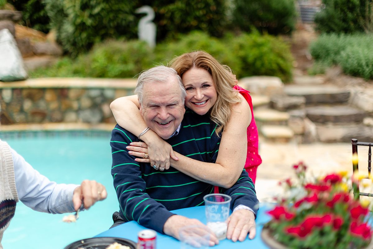 A guest in a bright pink dress leaning down to wrap her arms around a laughing older man in a striped sweater at the poolside table