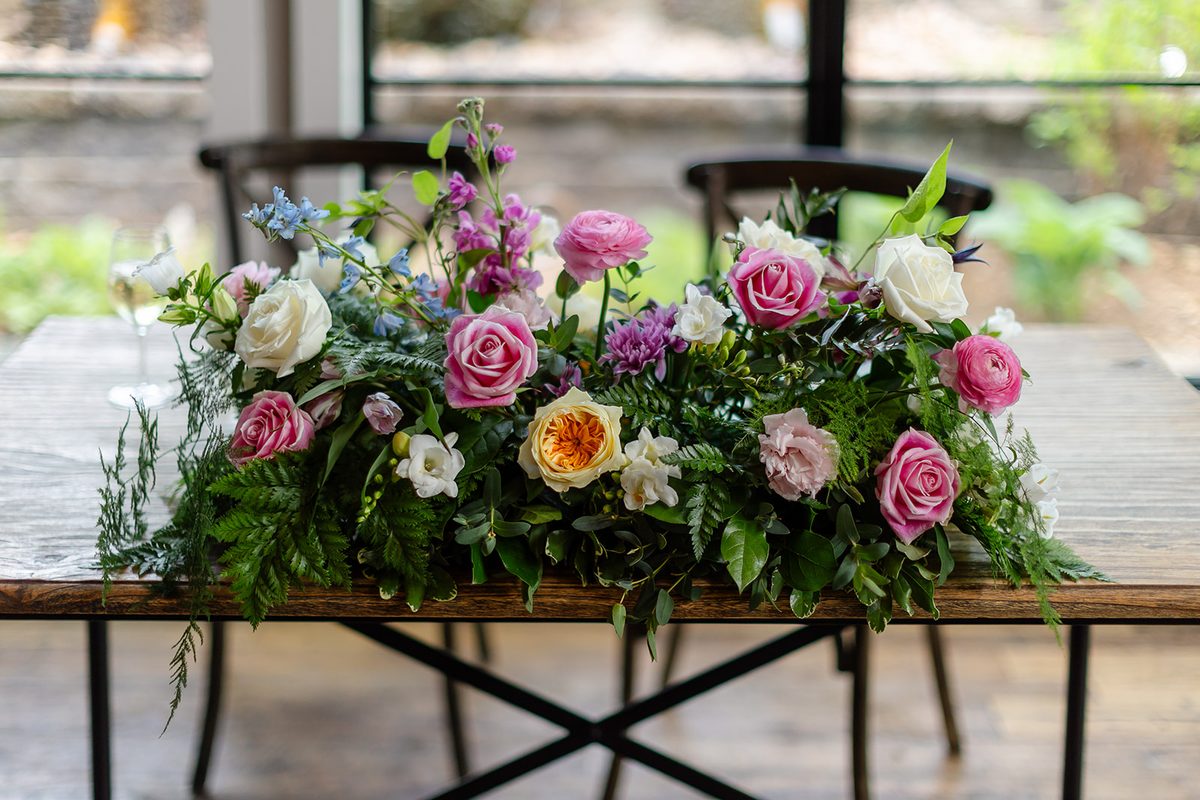 A long low arrangement of pink and cream roses, ranunculus, purple mums, and ferns running down the center of the couple's wooden table by the window