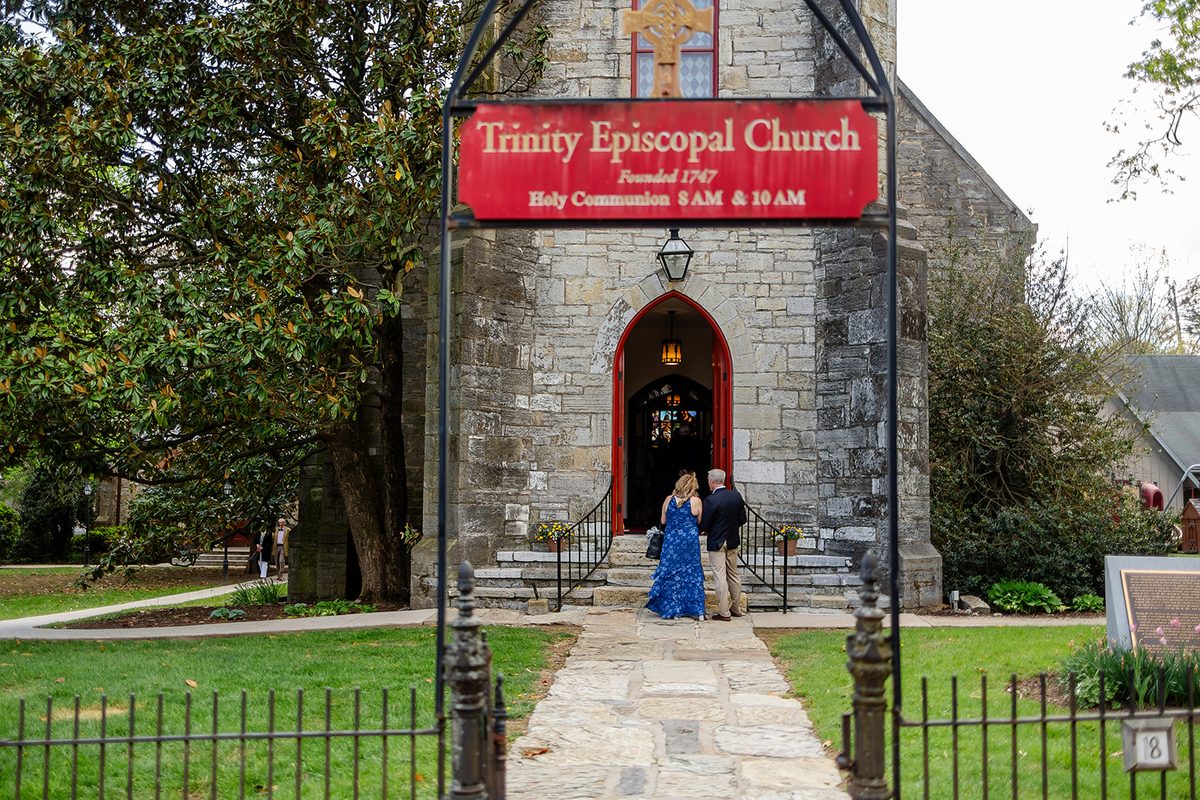 The stone front of Trinity Episcopal Church in Shepherdstown with its red doors open, the parish sign reading Founded 1747 as guests arrive