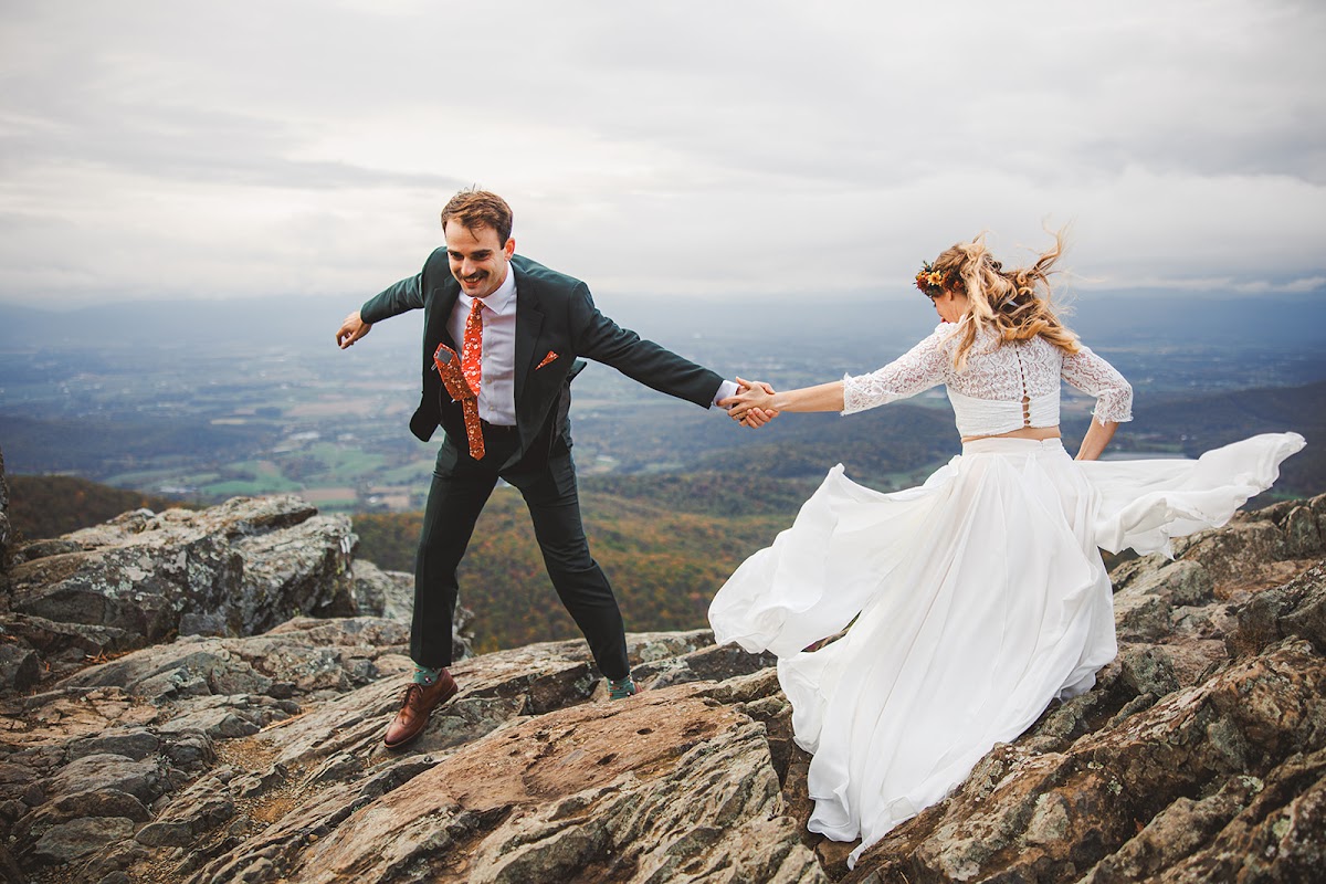 A groom leading his bride across a windy summit boulder, her skirt streaming out behind her