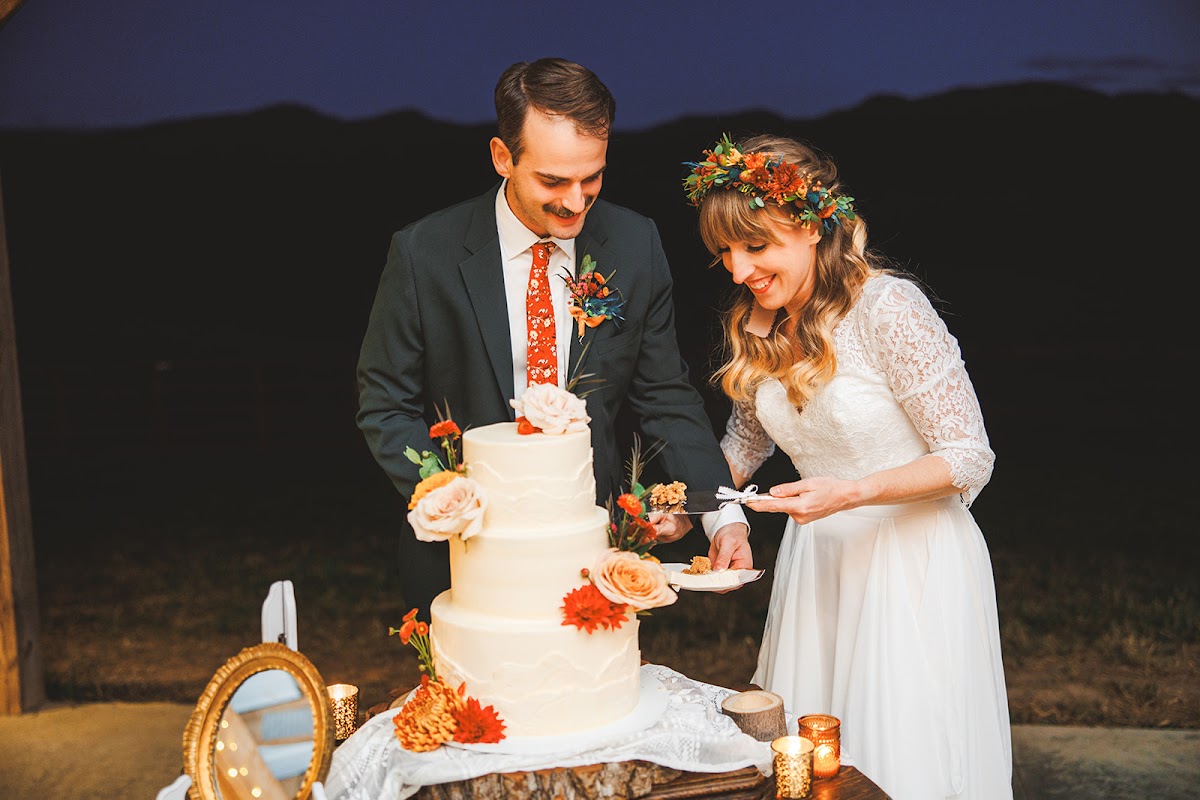 Sara and Ryan cutting the cake together at dusk, candles glowing on the table and the dark ridge of the Blue Ridge behind them