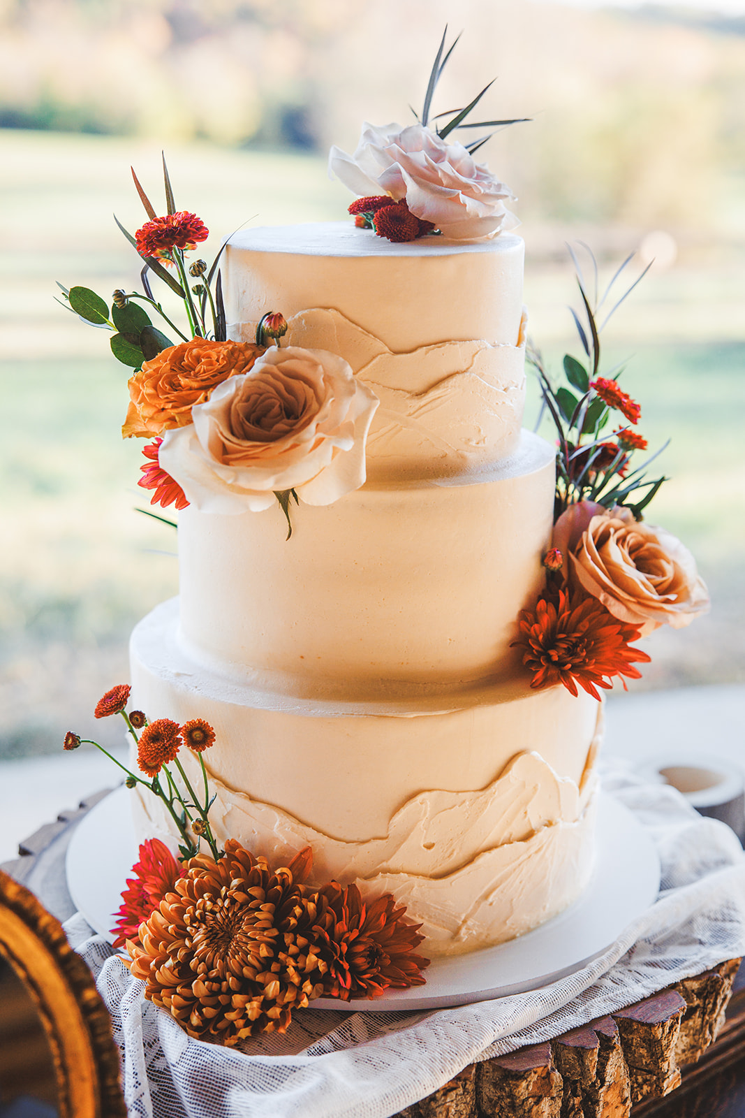 Two-tier white wedding cake with textured mountain-ridge icing, decorated with orange and peach roses and mums, photographed outdoors with the green valley behind