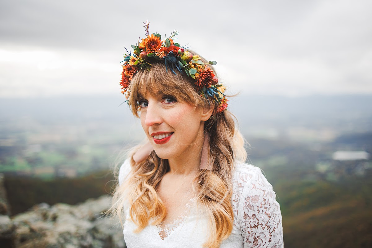 Portrait of Sara smiling in her orange, rust, and blue flower crown on the overlook, the valley towns soft and far below