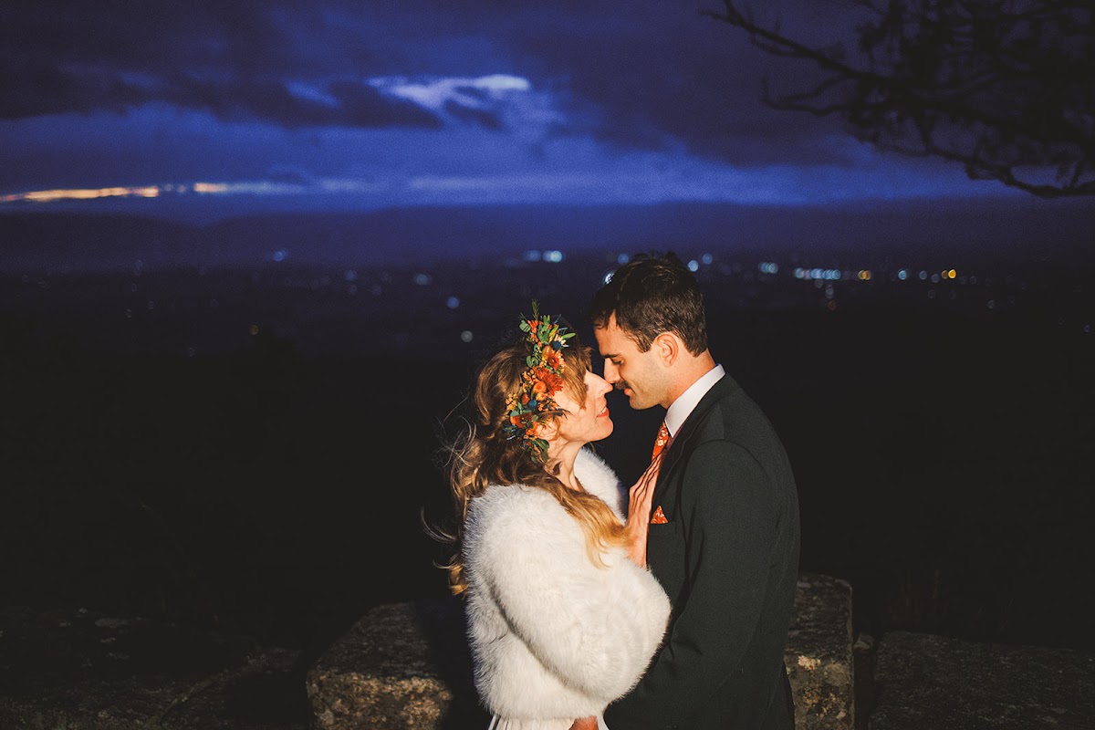 Close night portrait of Sara and Ryan touching foreheads, her white fur wrap and flower crown lit warm against the deep blue sky and distant valley lights