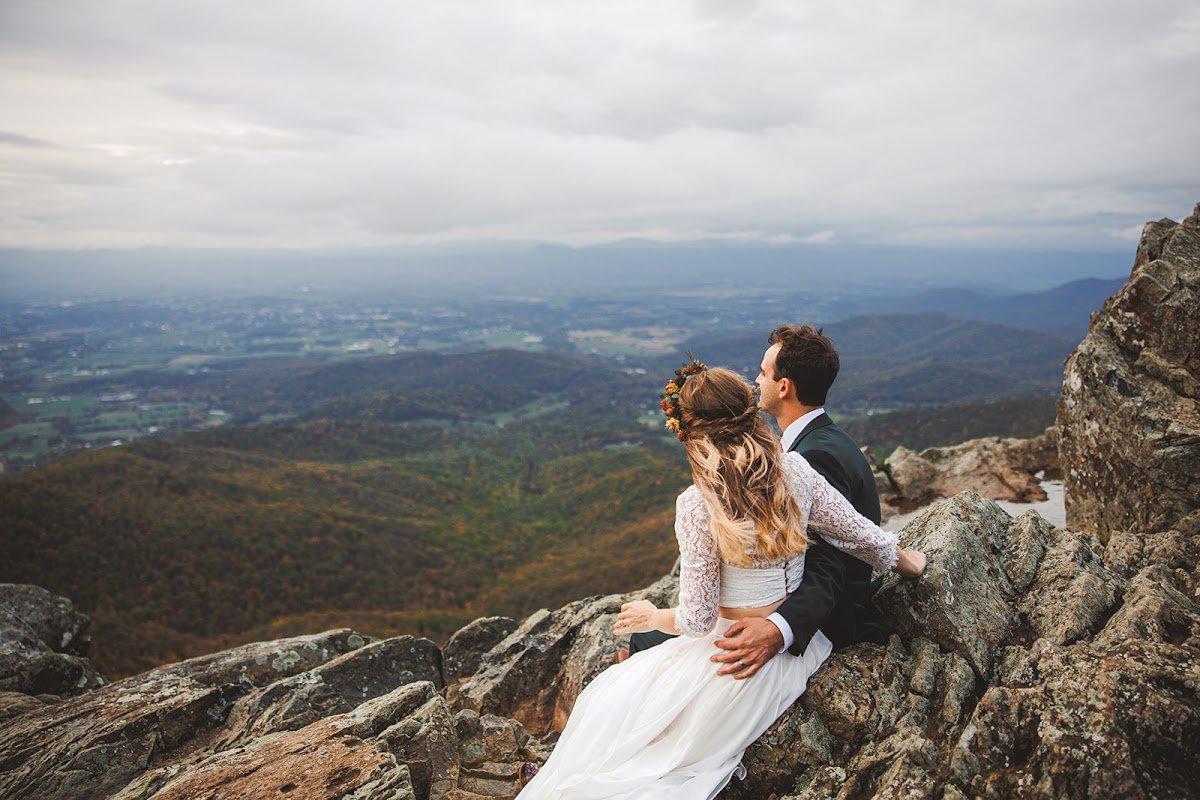 Sara and Ryan seated together on a rocky outcrop, seen from behind, looking out over the wide valley and layered Blue Ridge mountains