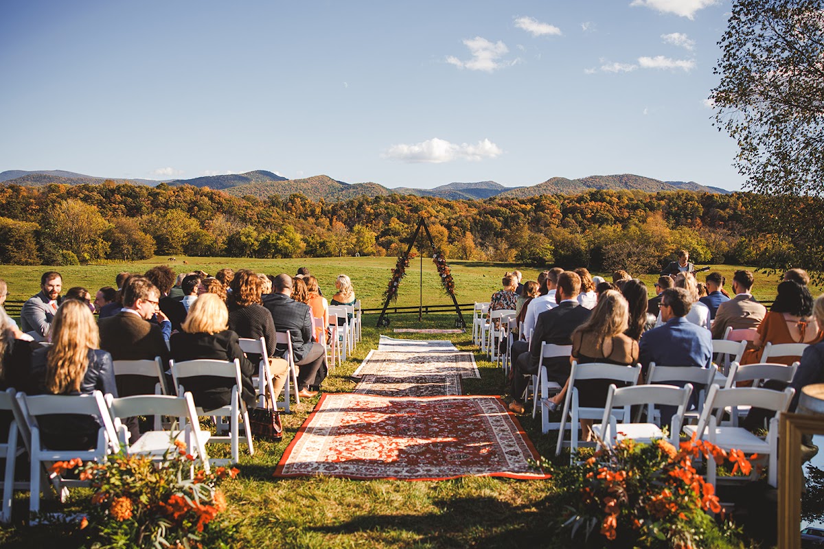 The ceremony at River's Bend Ranch seen from behind the seated guests: a rug-lined aisle with orange florals leading to a triangle arch, autumn mountains beyond