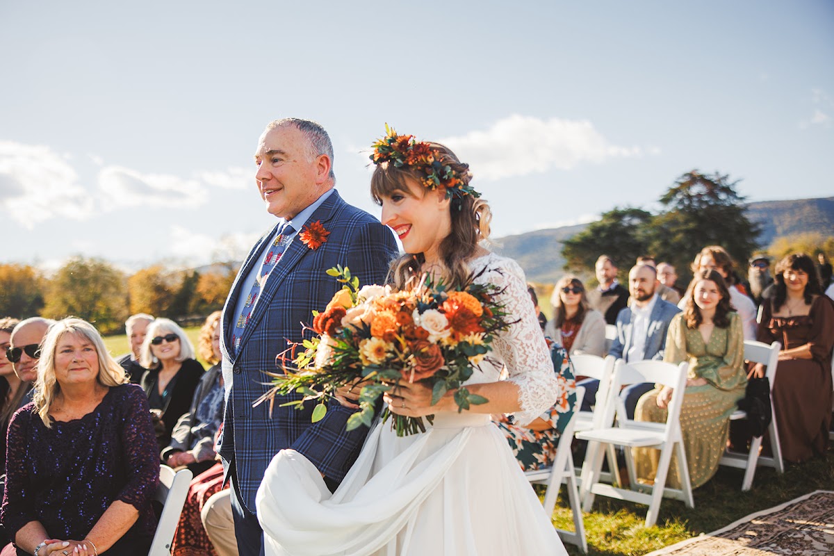 Sara walking down the aisle at River's Bend Ranch in her flower crown, carrying an orange and rust bouquet, escorted past seated guests in the autumn sun