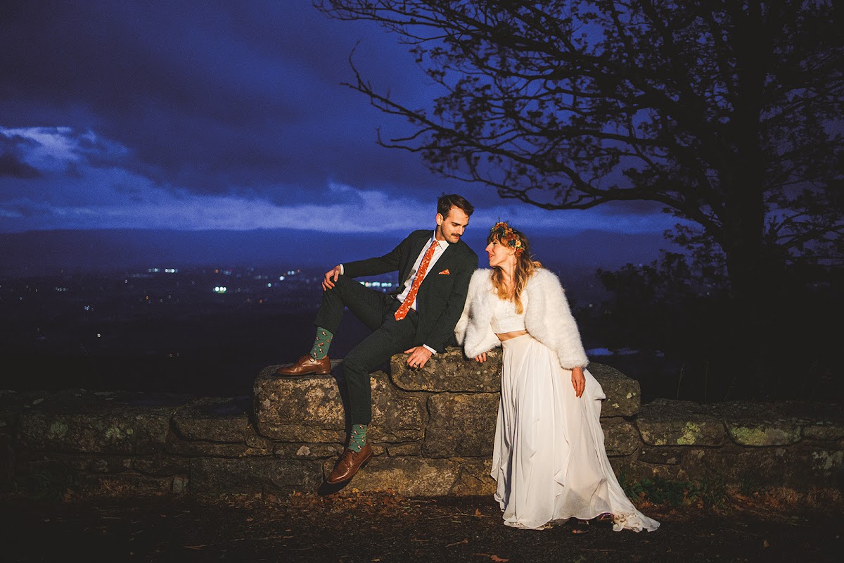 A couple resting on a stone wall at blue hour with town lights scattered far below