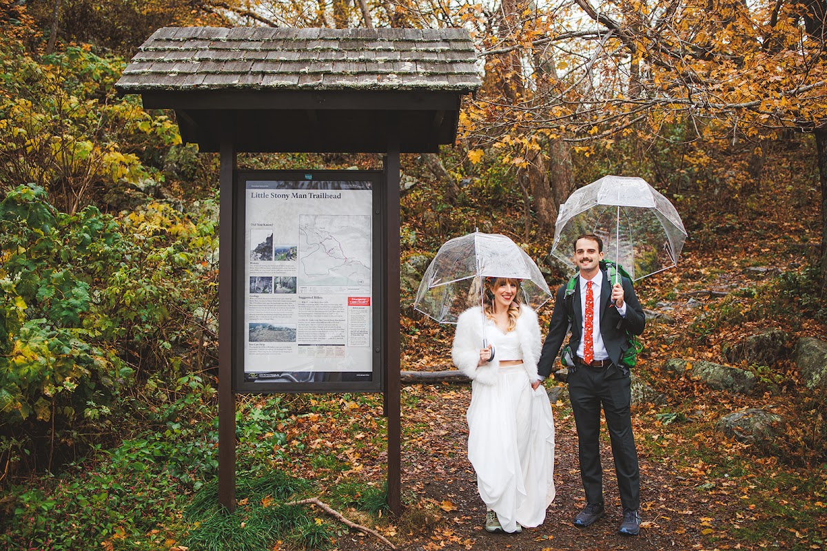 Sara and Ryan standing at the Little Stony Man Trailhead sign with clear umbrellas, Sara in a white fur jacket and Ryan carrying a green pack over his suit