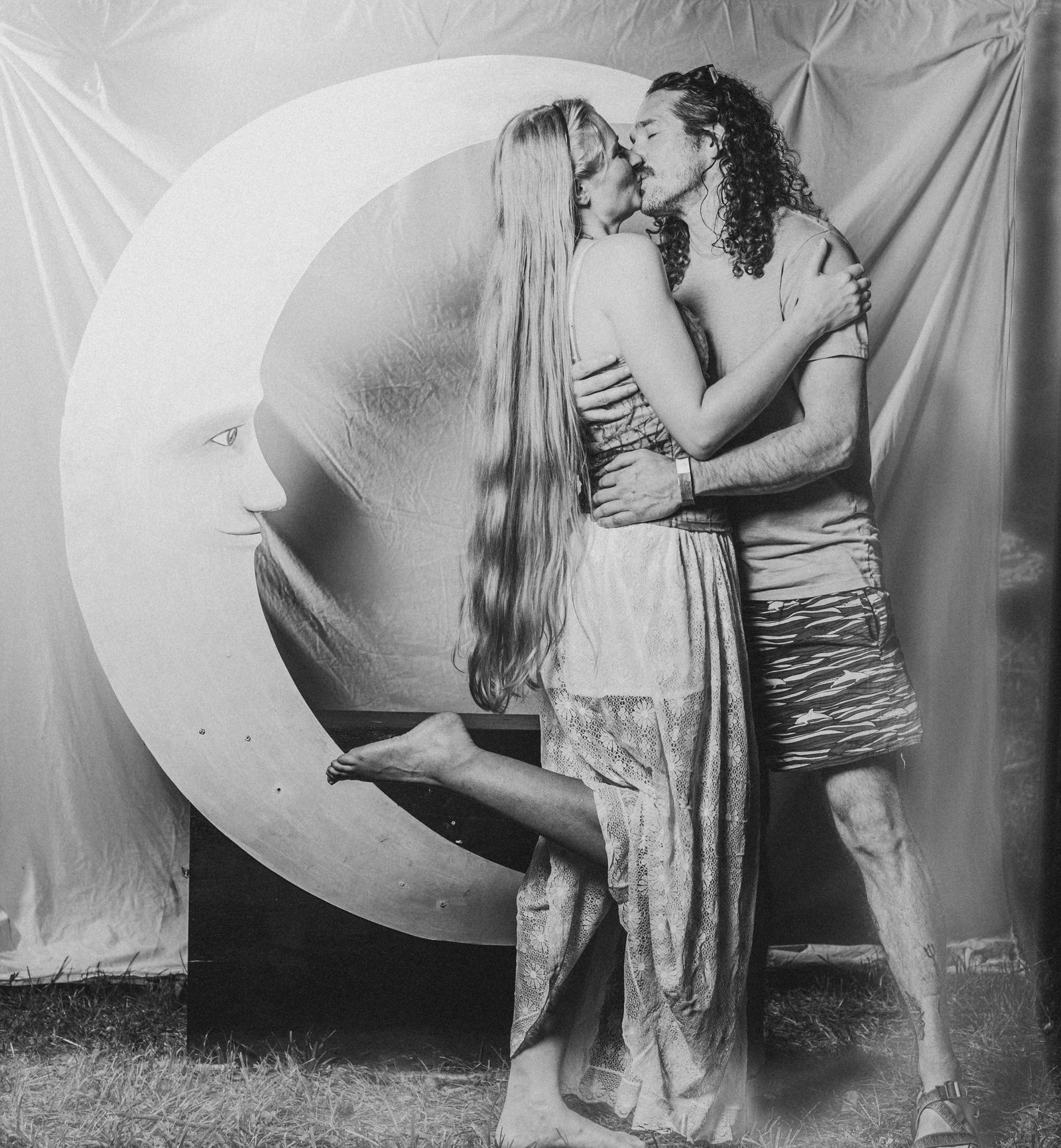 A couple kissing against the big crescent moon at the Tomato Jam Moon Booth, her bare foot kicked up behind her, in black and white
