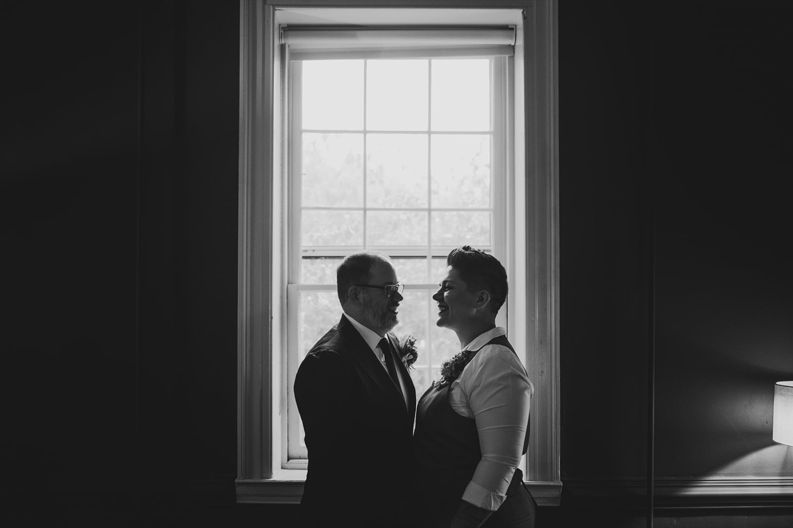 Lauren and Bert laughing together beside a tall window, in black and white