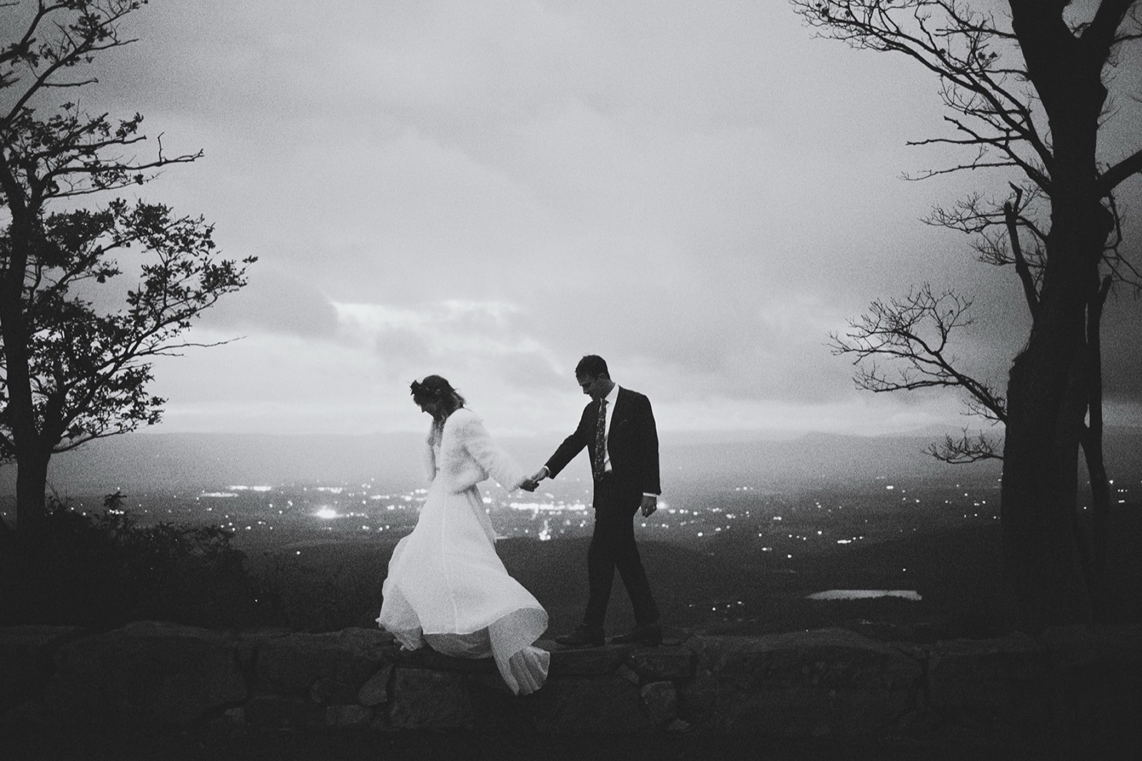 Sara and Ryan stepping along a stone wall above the valley lights at dusk, in black and white