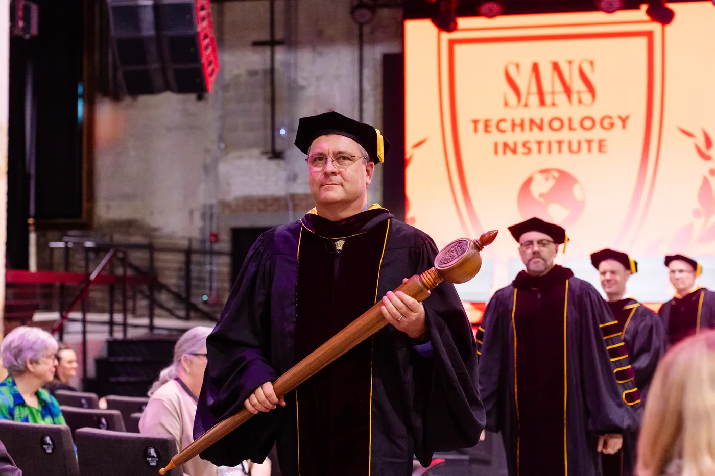 A faculty member in academic robes stands in front of a SANS Technology Institute commencement banner.