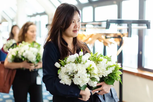 Woman carrying white floral arrangement in a sunlit corridor.