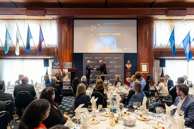 Wide view of a dinner ballroom with a handshake on stage and the audience at round tables.