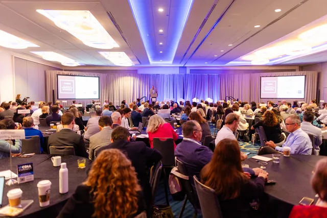 Full ballroom of attendees, blue ceiling glow.