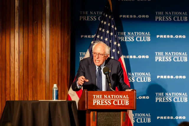 Bernie Sanders at the National Press Club podium beside the American flag and the blue press backdrop.