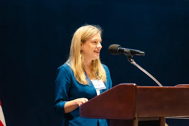Woman speaking at a wooden podium against a navy curtain.
