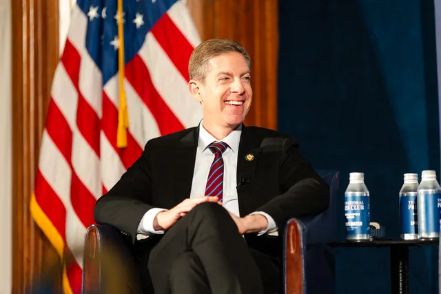 Man laughing in an armchair beside the American flag.