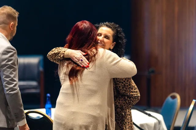 Two attendees hug warmly, with flags and the room behind them.